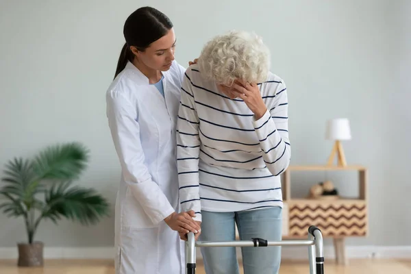 Young woman physiotherapist caregiver consoling sad injured old grandma patient