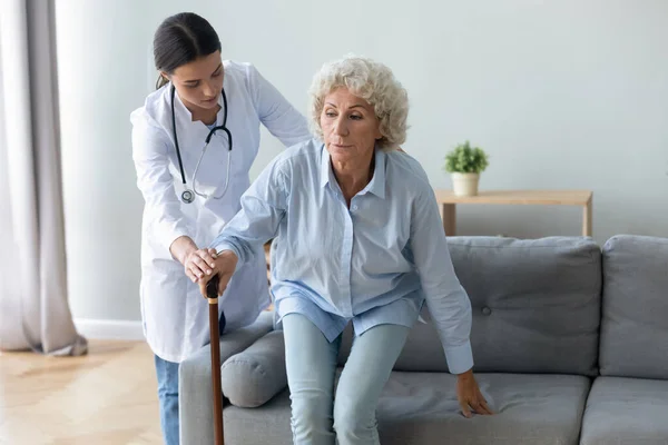 Sjuksköterska hjälp mormor få upp från soffan håll promenadkäpp — Stockfoto