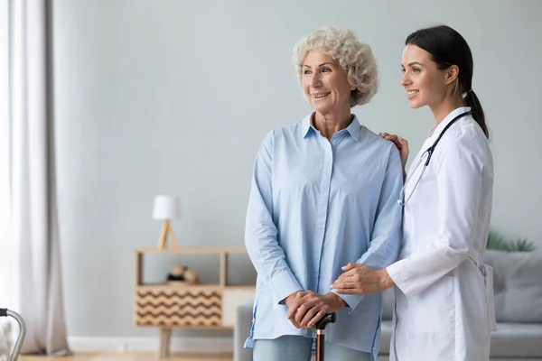 Glad kvinnlig läkare hjälpa mormor hålla promenadkäpp tittar bort — Stockfoto