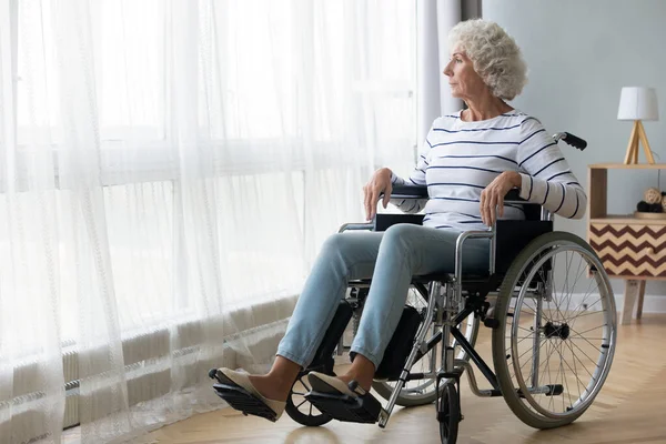 Pensivo deficiente avó sênior sentar-se em cadeira de rodas olhar através da janela — Fotografia de Stock