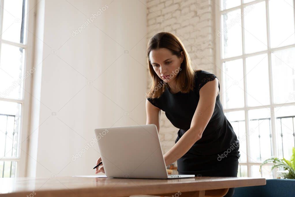 Worried young businesswoman leaning over table, typing answer to email.