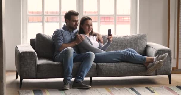 Relaxado casal milenar usando telefone levando selfie lounge no sofá — Vídeo de Stock