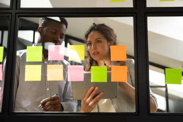 Koncentrerad blandras unga kollegor står nära glas kanban styrelse. — Stockfoto