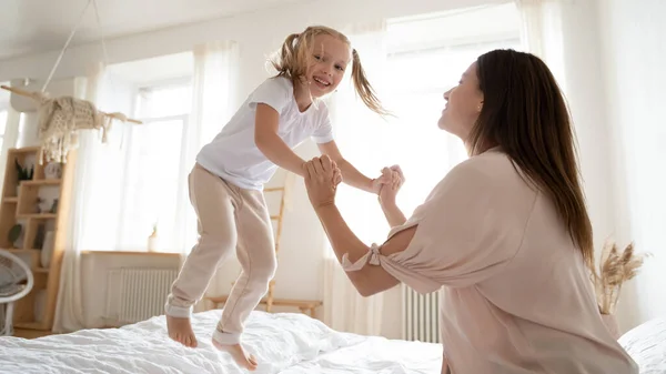 Llena de alegría preescolar adorable hija saltando en la cama, sosteniendo las manos de mamá . —  Fotos de Stock