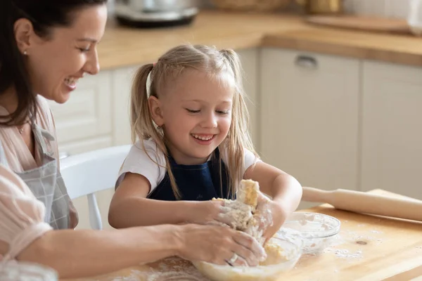Adorável filha ajudando sorrindo jovem mãe amassar massa de farinha por mãos . — Fotografia de Stock