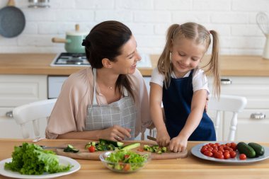 Mutlu genç anne, küçük anaokulu kızının sebze kesişini izliyor..
