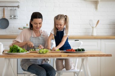 Small cute daughter helping young proud mommy cutting fresh vegetables. clipart