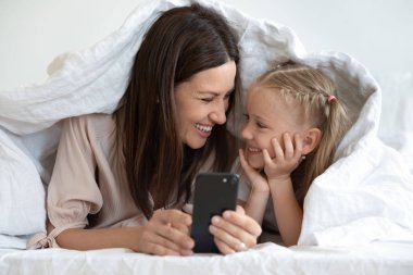 Mutlu anne, kızıyla battaniyenin altında uzanıyor, akıllı telefon kullanıyor..