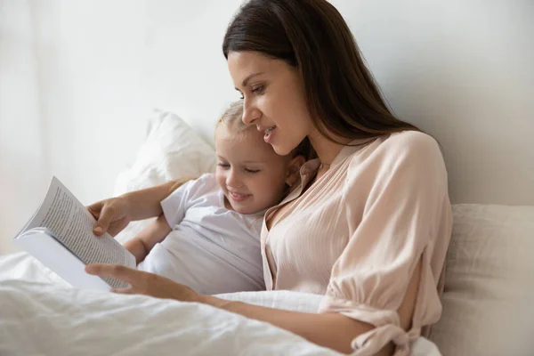 Young mommy lying in bed with daughter, reading book. — Stock Photo, Image