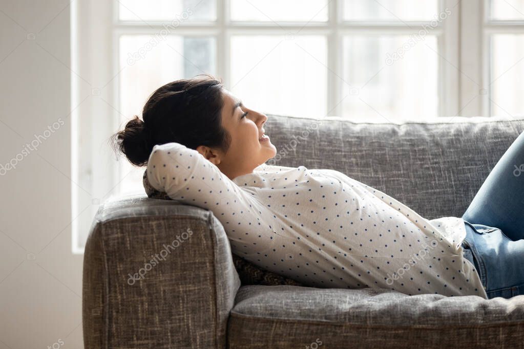 Happy millennial tranquil indian woman lying on comfortable couch.