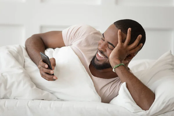 Happy african laki-laki Amerika berbaring di tempat tidur menggunakan ponsel — Stok Foto