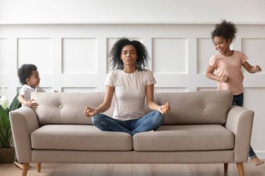 Gürültücü çocuklar koşuştururken Afrikalı anne kanepede meditasyon yapıyor.
