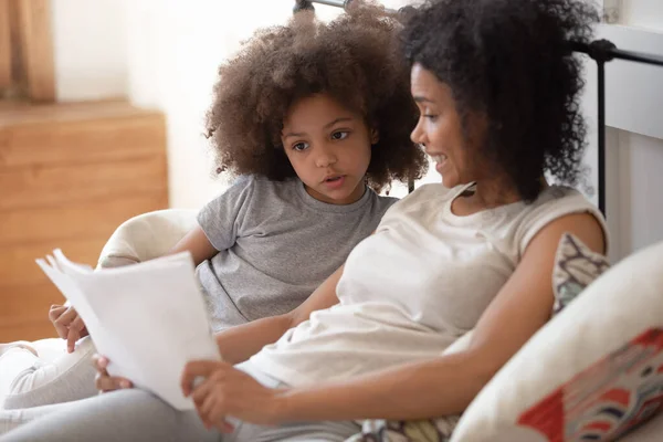 Çift ırklı anne, küçük kızıyla birlikte yatak odasında kitap okuyor. — Stok fotoğraf