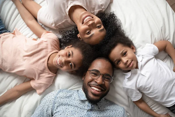 Χαρούμενη αφρικάνικη οικογένεια ξαπλωμένη στο κρεβάτι χαμογελώντας κοιτάζοντας την κάμερα — Φωτογραφία Αρχείου