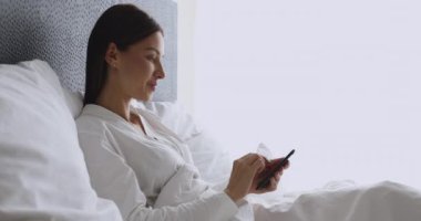 Mutlu genç bayan sabahları yatağında telefon kullanıyor.