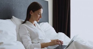 Dizüstü bilgisayar kullanan neşeli genç kadın zaferi kutluyor. Yatağa otur.