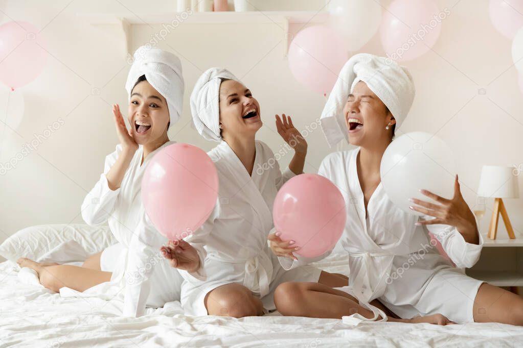 Excited multiethnic young ladies sit on bed with pink balloons