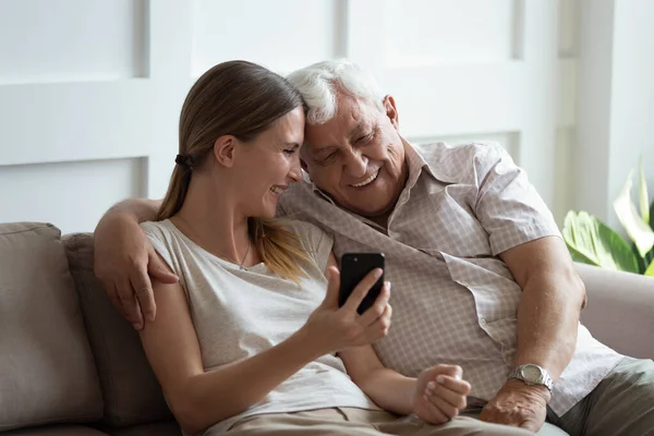 Szczęśliwa młoda kobieta pokazuje śmieszne zdjęcia na smartfonie do ojca. — Zdjęcie stockowe