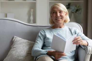 Kanepede oturan yaşlı bir kadın elinde kitapla kameraya bakıyor.