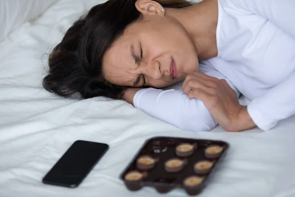 Mujer deprimida acostada en la cama cerca de celulares y dulces —  Fotos de Stock