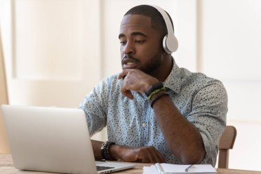 Kulaklıklı konsantre olmuş çift ırklı adam dizüstü bilgisayarda Webinar izliyor.