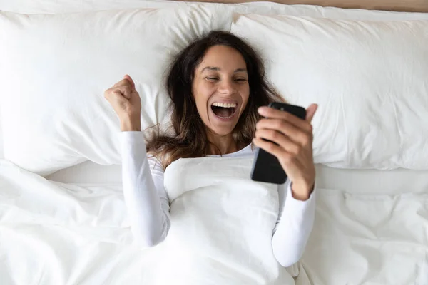 Woman lies in bed holding smartphone scream with joy — Stock Photo, Image