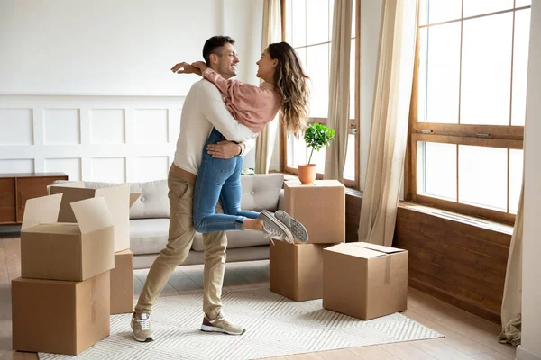 Happy husband lifting excited wife celebrating moving day with boxes — Stock Photo, Image