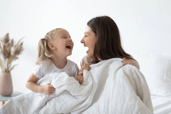 Happy mother laughing with kid daughter holding duvet in bed