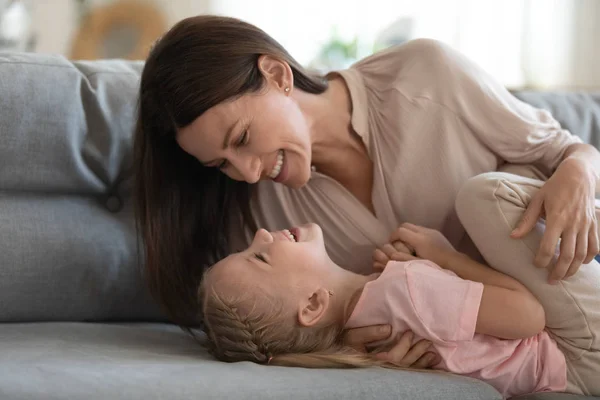Sonriente madre e hija pequeña jugando cosquillas en el sofá —  Fotos de Stock