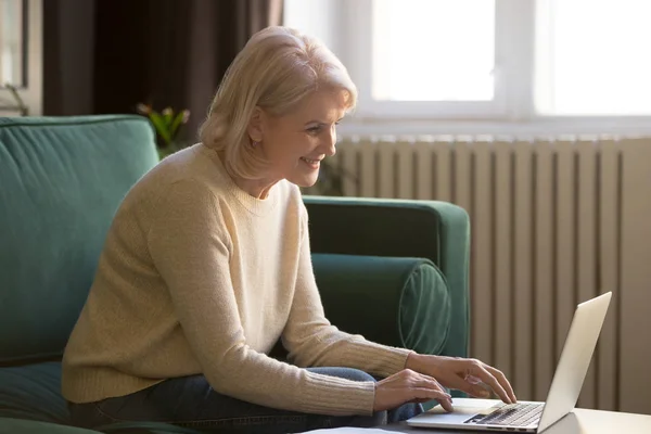 Ältere Frau sitzt auf Couch und tippt am Computer — Stockfoto