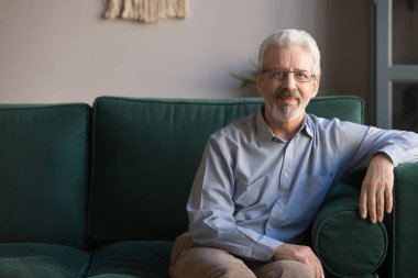 Kanepede oturmuş kameraya bakan yakışıklı, yaşlı bir adam.