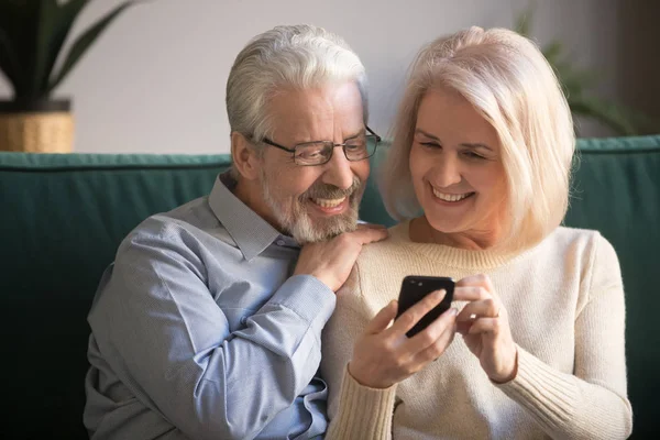 Mature attractive couple sitting on couch using smartphone — Stock Photo, Image