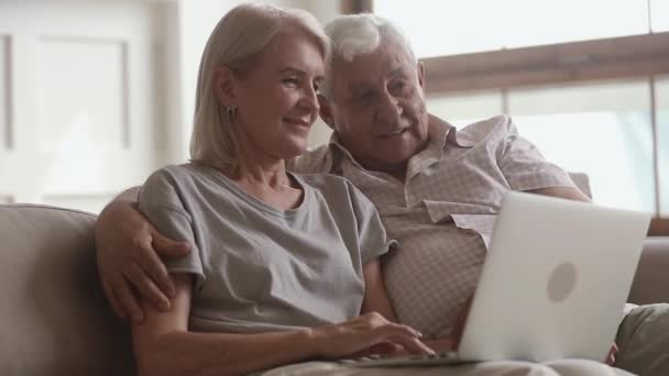 Счастливая пожилая пара разговаривает смеясь обнимаясь, используя ноутбук вместе — стоковое видео