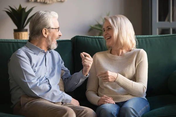 Cheerful elderly spouses sitting on couch chatting feel happy