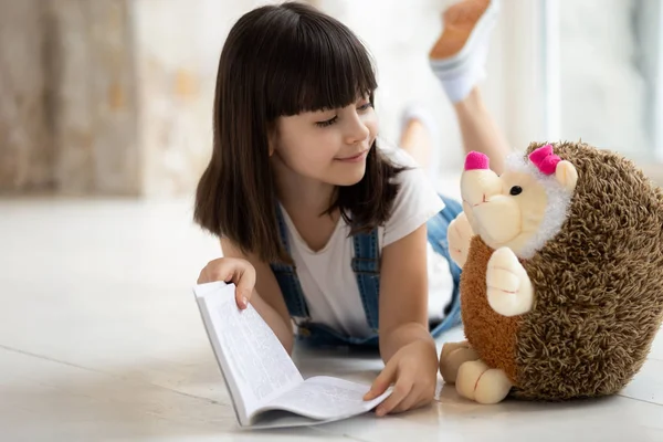 Cute small girl reading book to toy hedgehog — Stock Photo, Image