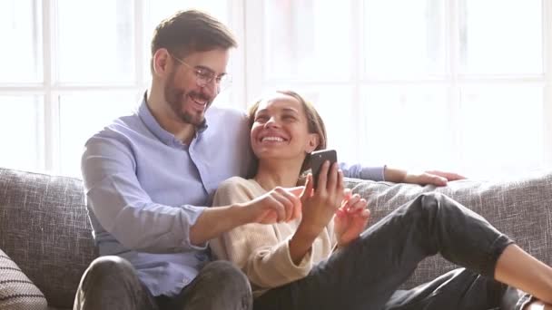 快乐的夫妻笑着看着智能手机坐在沙发上 — 图库视频影像