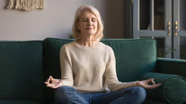 Serene elderly woman sit in lotus position meditating at home