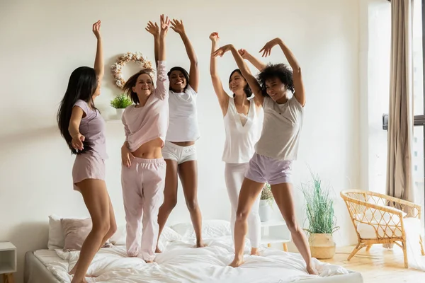 Glückliche multiethnische Frauen tragen Pyjamas, die gemeinsam auf dem Bett tanzen — Stockfoto