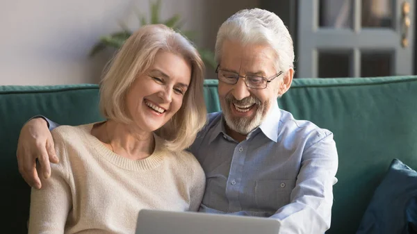 Cheerful senior couple sitting on couch watching movie on laptop