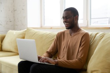 Gülümseyen Afrikalı Amerikalı adam oturma odasında dizüstü bilgisayar kullanıyor.