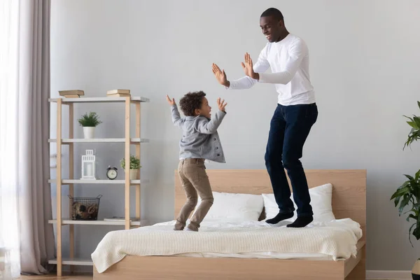 Joven afroamericano alegre saltando en la cama con su hijo . —  Fotos de Stock