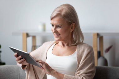 50 'li yaşlarda bir kadın kanepeye oturmuş tablet bilgisayar kullanıyor.