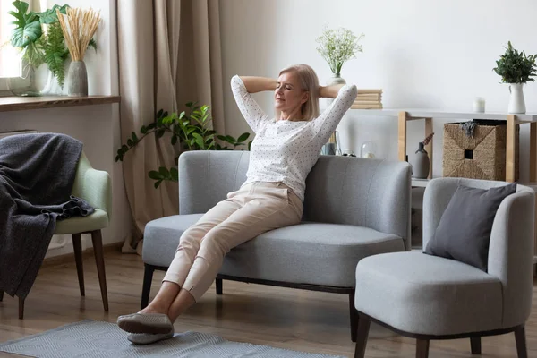 Attractive elderly woman resting on couch putting hands behind head
