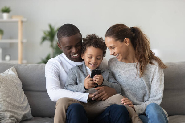 Happy black husband holding son on knees, playing on phone.