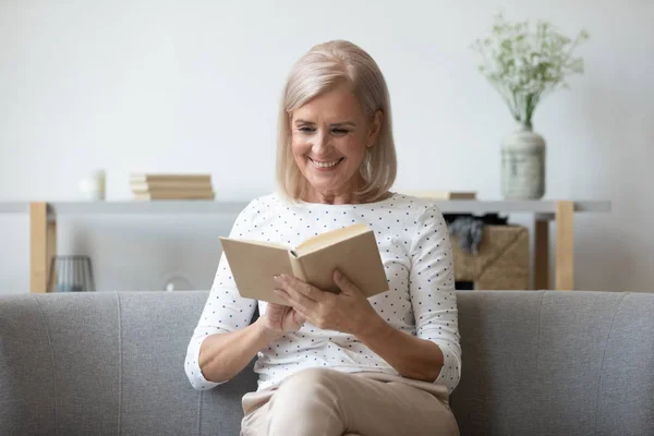 Attractive elderly female resting on couch reading book enjoy weekend