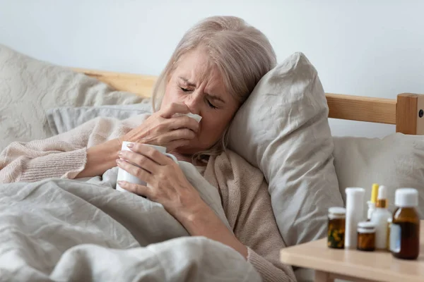 Elderly woman lying in bed blowing runny nose in handkerchief — Stock Photo, Image