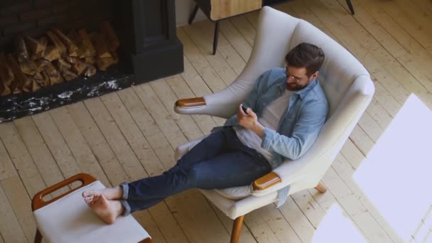 Happy man zitten op comfortabele stoel in huis met behulp van telefoon — Stockvideo