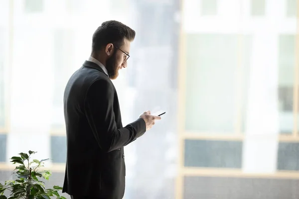 Erfolgreicher Geschäftsmann steht in der Nähe von Fenstern und surft mit dem Handy — Stockfoto