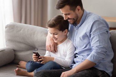 Baba küçük oğlunu kucaklıyor. Akıllı telefon kullanarak birlikte kanepede oturuyor.