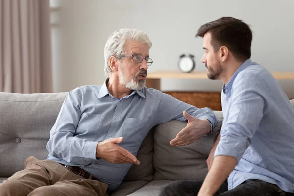 Serious elderly father and grownup son sitting on couch talking — Stock Photo, Image